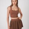 A young woman with long, straight hair wearing a brown sleeveless crop top and a matching pleated skirt, standing against a plain light gray background.